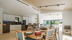 Eine moderne Küche mit weißen Schränken, ein Essbereich mit einem Holztisch und Stühlen sowie ein Blick ins Freie durch große Fenster.