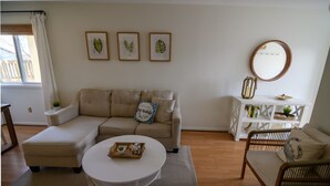 Ein Wohnzimmer mit einem beigefarbenen Sofa, einem weißen Couchtisch und einem Korbstuhl. An der Wand befinden sich gerahmte Blätter-Kunstwerke, ein runder Spiegel und eine weiße Konsole mit Büchern und Dekoration.