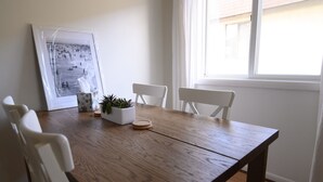Ein Esstisch mit Holzplatte, umgeben von weißen Stühlen. An der Wand hängt ein gerahmtes Foto, und auf dem Tisch steht ein Topfpflanze.