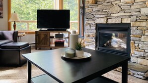 Ein Wohnzimmer mit Kamin aus Stein, einem Fernseher auf einem Holzständer und einem großen Fenster mit Blick auf Bäume.