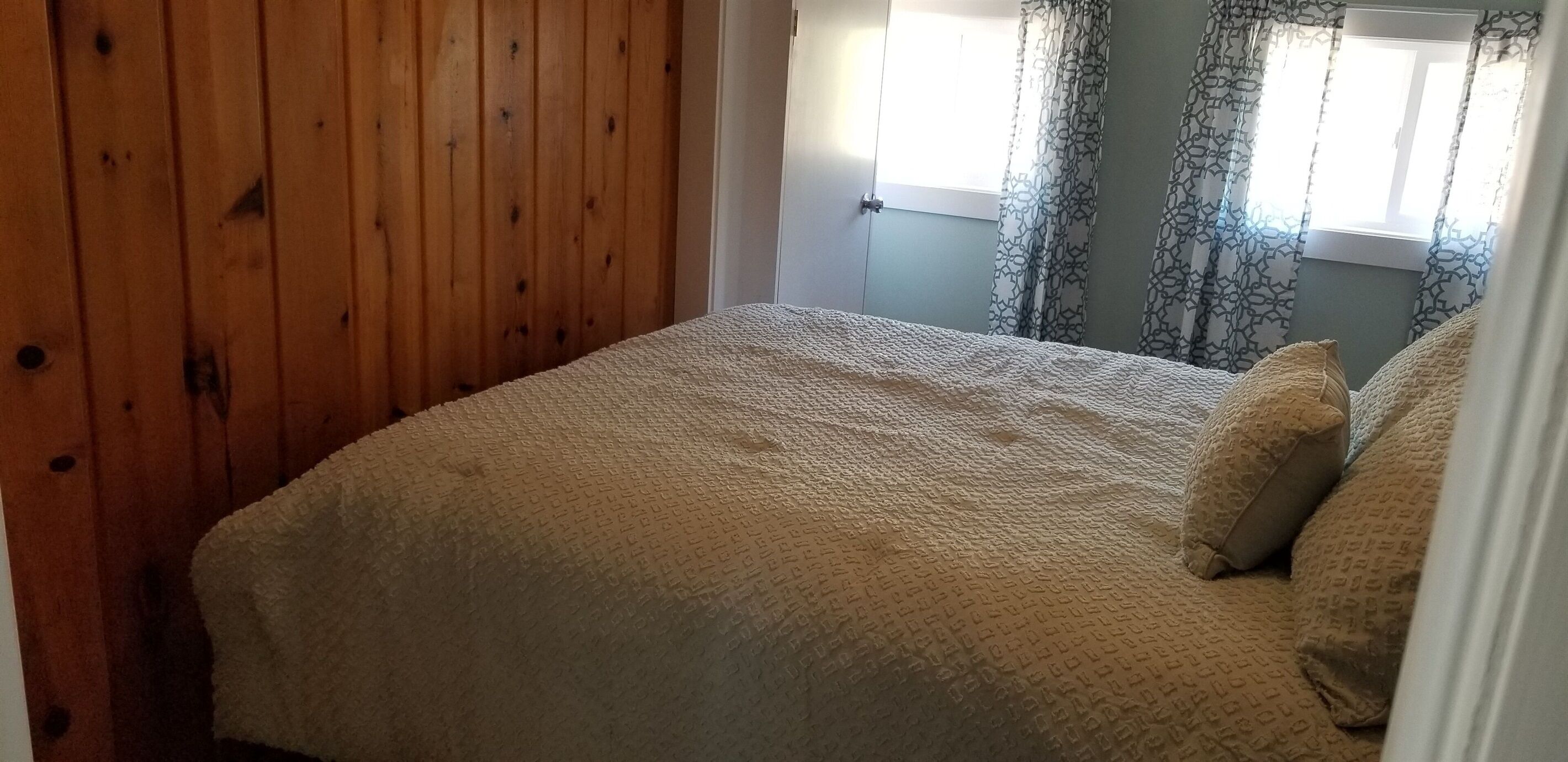 Ett sovrum med en väggpanel av trä, en säng med ett mönstrat överkast, draperade gardiner och ett fönster med utsikt.