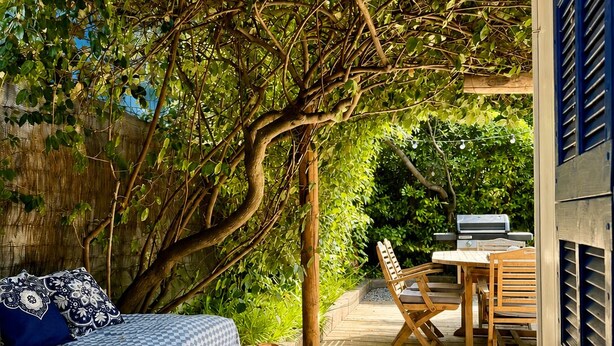 A wooden deck with a bed and a table with chairs, surrounded by lush greenery and trees.