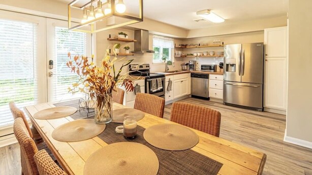 Una cocina moderna con armarios blancos, electrodomésticos de acero inoxidable y un comedor con mesa de madera y sillas de mimbre.
