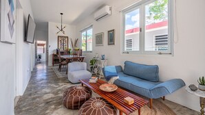 A modern living room with a blue sofa, wooden coffee table, and decorative pillows. There is a dining area with a table and chairs, and a kitchen in the background.