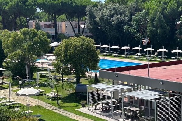 A pool area with sun loungers and umbrellas, surrounded by trees and a building with a red roof.