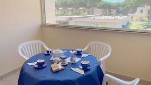 Un coin repas avec une table dressée pour deux personnes, agrémenté d’une nappe bleue, de chaises blanches et offrant une vue sur les arbres et les bâtiments à travers une fenêtre.