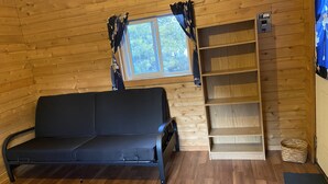L’intérieur d’un chalet en bois, avec un canapé noir, une fenêtre avec des rideaux et une étagère en bois.