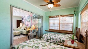 A room with two beds, a ceiling fan, a window with blinds, and a view of a dining area.