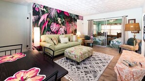 A room with a green sofa, a dining table, a patterned rug, and a tropical leaf mural on the wall.