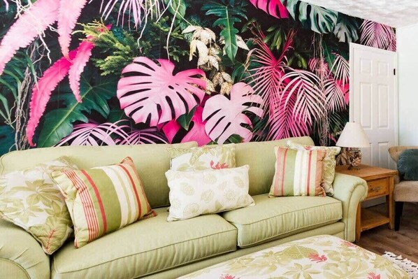 A living room with a green sofa, patterned cushions, a wooden side table, and a tropical-themed mural on the wall.
