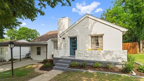 A single-story house with a white exterior, a blue front door, and a small front yard with steps leading to the door. - Spacious 5BR Near Bishop Arts - 5 Min Drive! (Dallas)