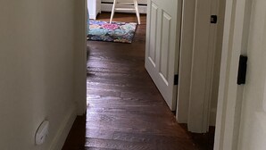 Um corredor com piso de madeira que leva a um quarto com porta branca, uma cadeira branca e um quadro na parede.