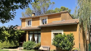 Una casa de dos pisos con techo de tejas, un porche con banca y un césped bien cuidado.