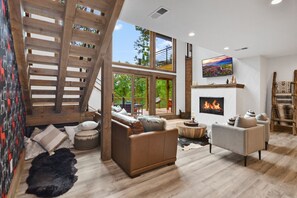 A modern living room with a fireplace, wooden staircase, and large windows. - Red Stag Ridge ~ Mtn Views ~ Hot Tub ~ EV Charger (Ellijay)