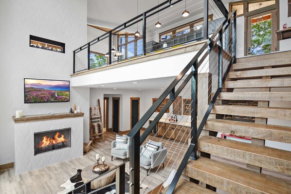 A spacious living area with a fireplace, a wooden staircase, and a balcony with hanging lights. - Red Stag Ridge ~ Mtn Views ~ Hot Tub ~ EV Charger (Ellijay)