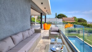 Un balcon extérieur moderne, doté d'un canapé gris, de meubles en osier et d'une balustrade en verre, offrant une vue sur une piscine et un espace verdoyant.