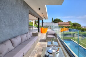 A modern outdoor balcony with a grey sofa, wicker furniture, and a glass railing overlooking a swimming pool and greenery. - Modern villa in Nice with sea view, private pool without view (Nice)