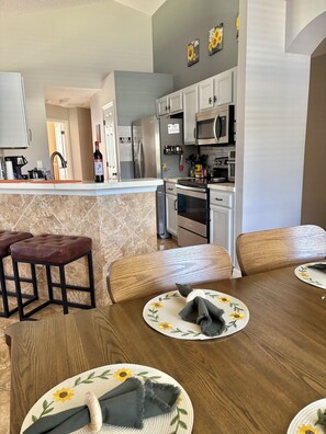 A modern kitchen with stainless steel appliances, a dining area with wooden chairs and a table set for two, and a bar area with stools.