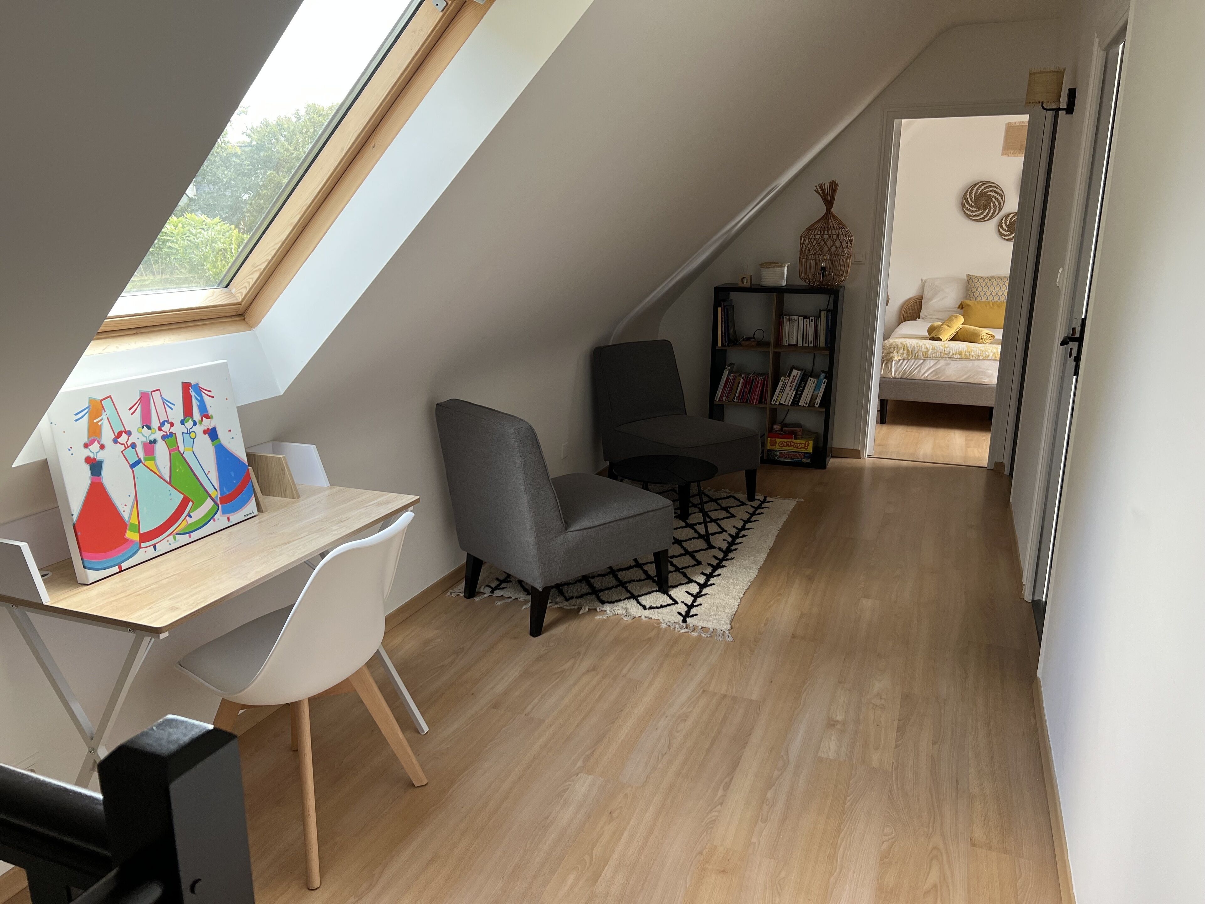 A modern loft bedroom with a sloped ceiling, a desk with a colorful painting, a gray armchair, a bookshelf, and a bed with yellow bedding.