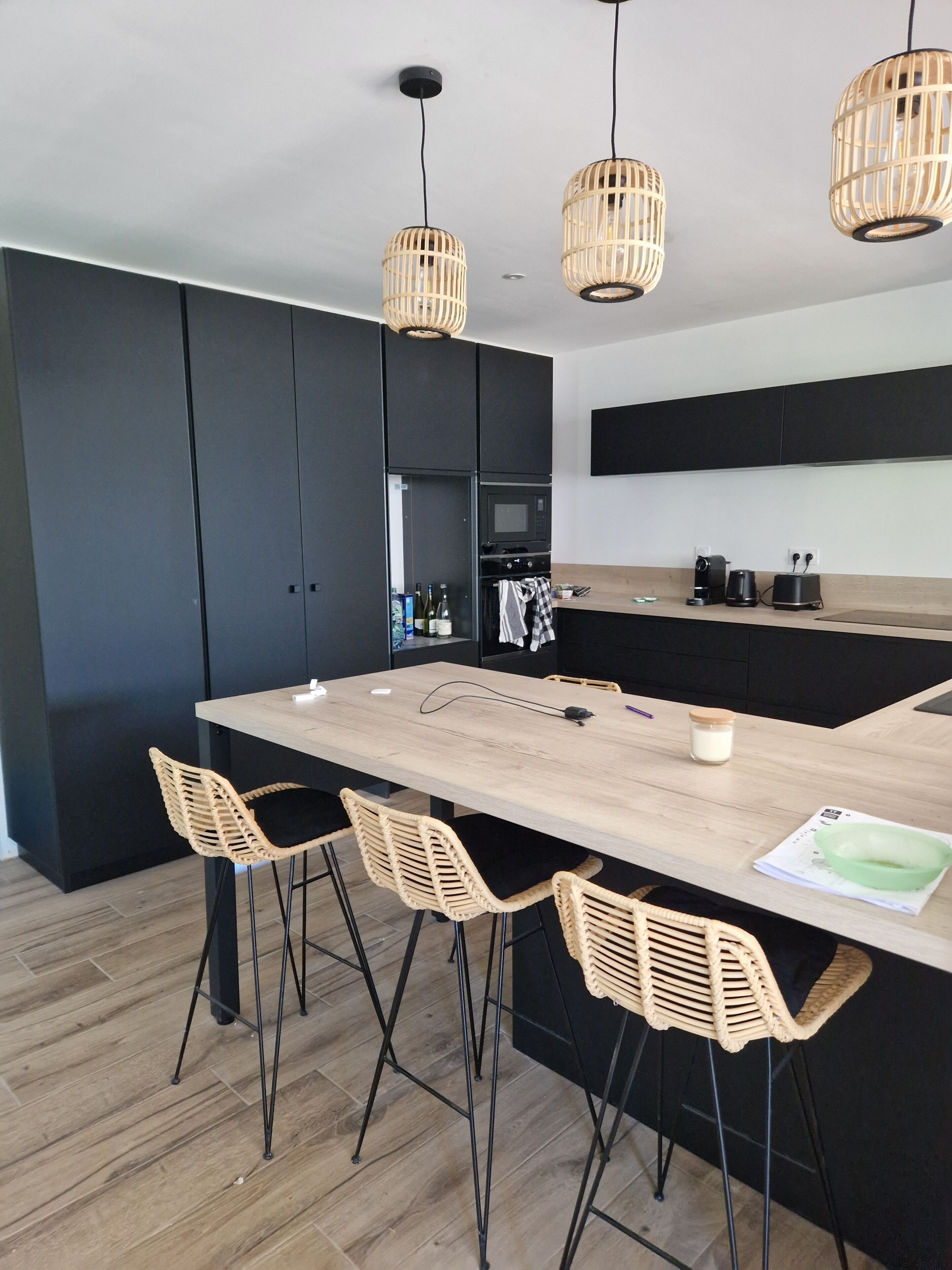 A modern kitchen with a central island, black cabinets, and wicker chairs.