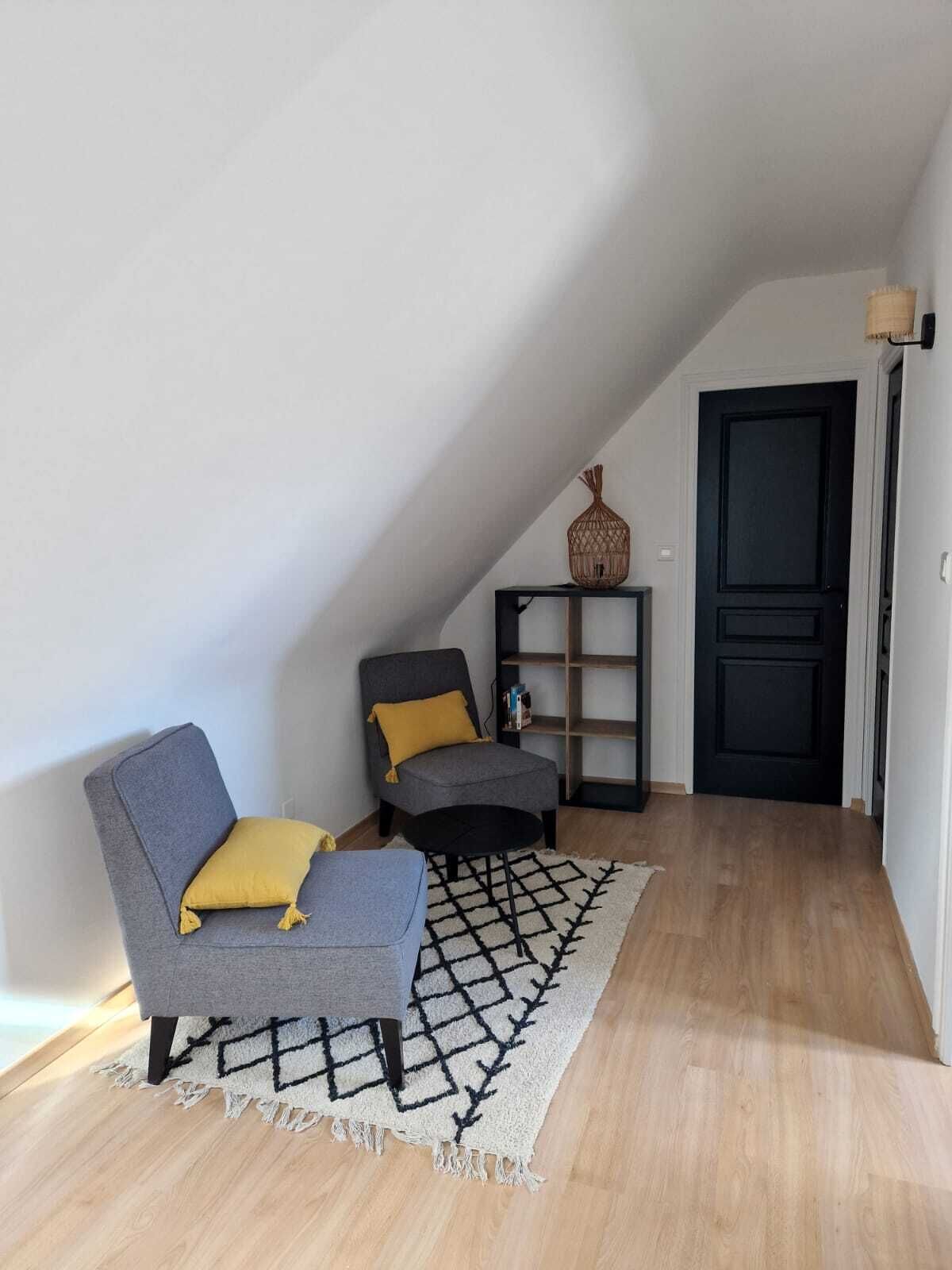 A cozy loft-style room with a sloped ceiling, two grey armchairs with yellow cushions, a black bookshelf, and a decorative woven basket.