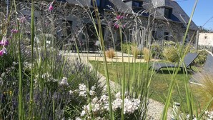 A stone house with a sloped roof, a garden with various plants, and a clear blue sky. - Des deux plages - PLERIN (PLERIN)