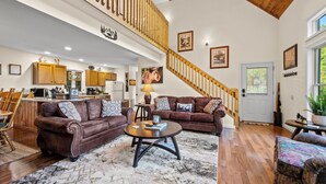 A spacious living room with a high ceiling, wooden staircase, and a kitchen in the background. - The Overlook /Hot Tub/Mountain Views/Babbling Creek/Fire Table (Maggie Valley)