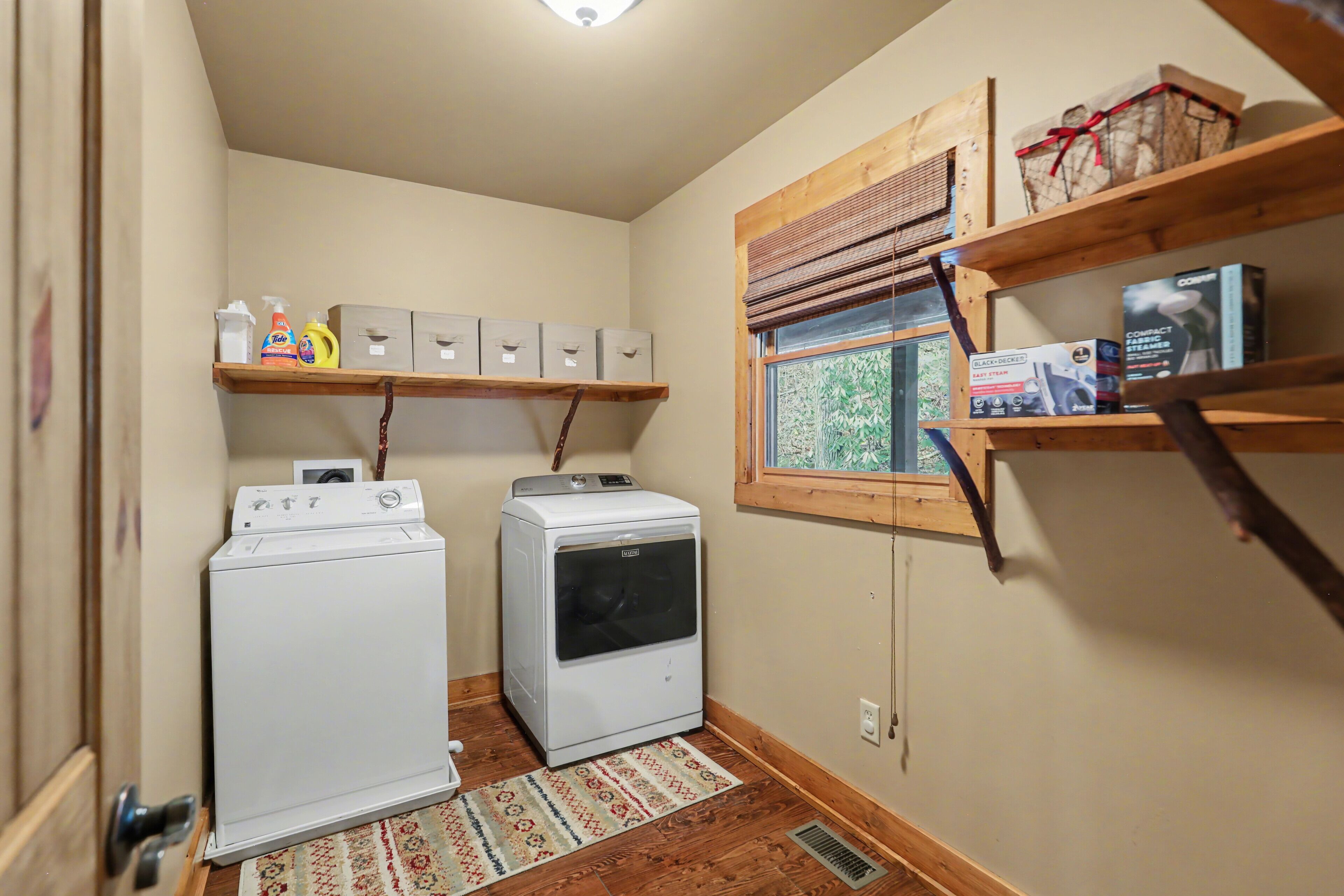 Un cuarto de lavado con dos lavadoras blancas, un estante de madera con productos de limpieza y una ventana con persianas.