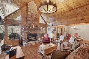 A cozy log cabin interior with a stone fireplace, wooden furniture, and large windows.