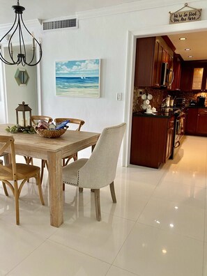 Un comedor con mesa y sillas de madera, una lámpara de araña, un cuadro de una playa y una cocina con armarios de madera y salpicadero azulejado.