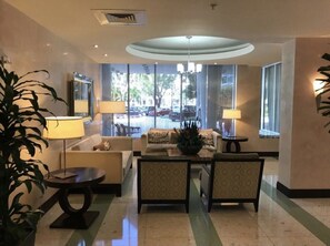 A hotel lobby with a reception desk, seating area, and large windows offering a view of the street. - Beautiful beach front condo with a magnificent beach and pool view. (Hollywood)