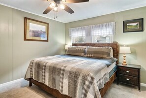 A bedroom with a wooden bed, a nightstand with a lamp, a ceiling fan, and two windows with curtains. - Gatlinburg. Downtown. 2br (Gatlinburg)