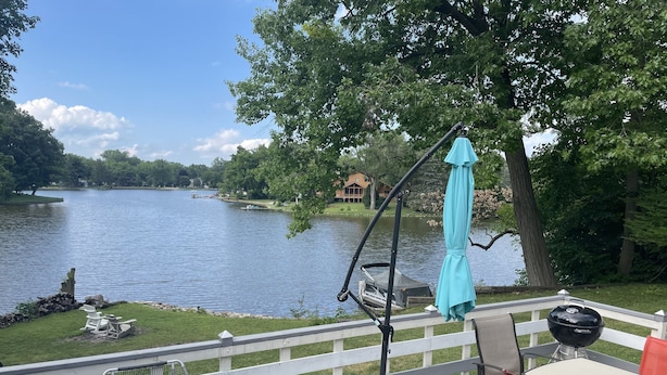Terraza con mesa y sillas con vista a un lago, una parrilla y un paraguas azul.