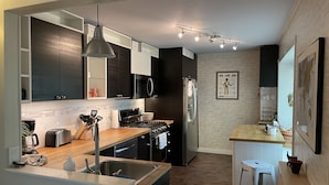 Una cocina moderna con armarios oscuros, un refrigerador de acero inoxidable, un microondas y una encimera de madera. Hay una pequeña isla con taburetes y una lámpara colgante arriba.