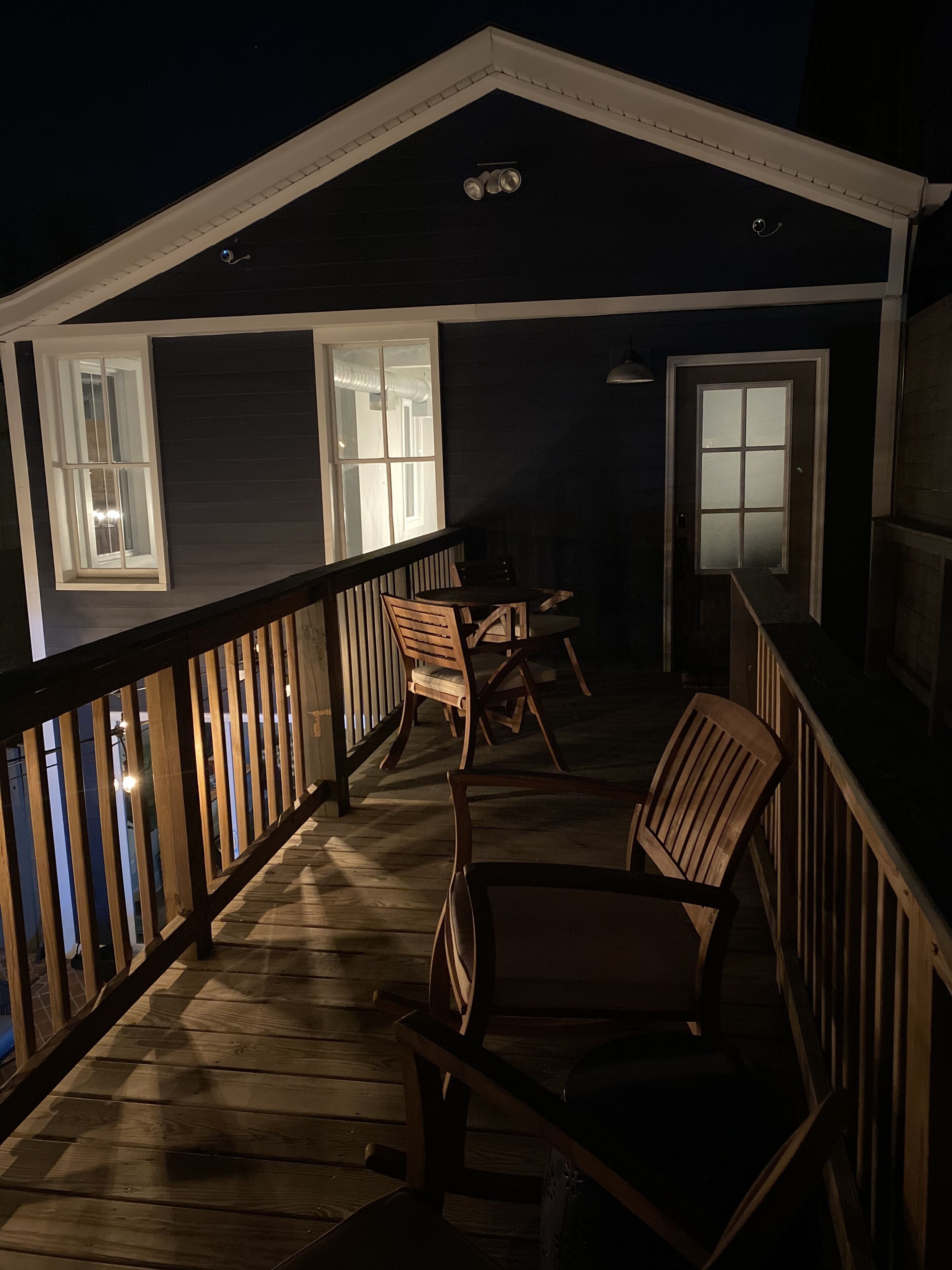 Una terraza techada al aire libre con sillas y una mesa de madera, barandilla y una puerta con paneles de vidrio esmerilado.