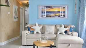 A living room with a beige sofa, starfish cushions, a glass dome with a plant, and a framed picture of a sunset over the ocean. - The Santi Paradise (Glen Burnie)