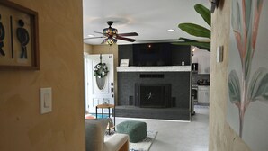 A modern living room with a fireplace, a ceiling fan, and a green ottoman. - The Santi Paradise (Glen Burnie)