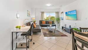 A modern living room with a sofa, coffee table, and a desk with a chair. There is a large window with curtains, a painting on the wall, and a view of buildings outside. - Stylish 2-Bedroom Apt, Free Parking, Walkable (Miami)