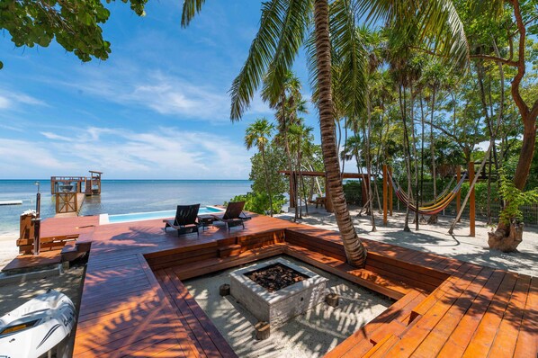 A wooden deck with a fire pit, lounge chairs, and a hammock overlooking a beach and ocean.