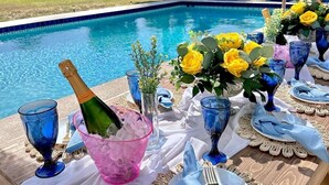 Montaje de comedor junto a la piscina con una mesa para dos, cristalería azul, un cubo de hielo rosa y rosas amarillas.