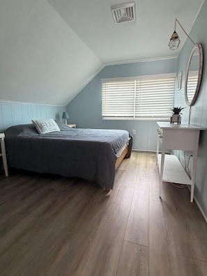 Une chambre à coucher comprenant un lit, une table de chevet, une commode et une fenêtre avec des stores.