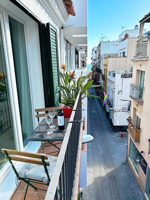 Een smalle stadstraat met gebouwen aan beide zijden, een balkon met een tafel, stoelen, een wijnfles en bloempotten.