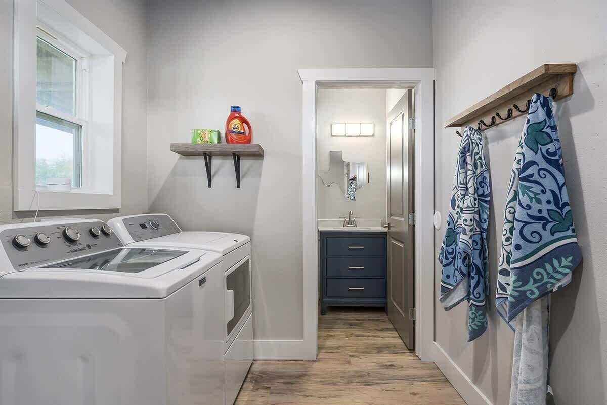 A laundry room with stacked washers and dryers, a shelf with cleaning supplies, and a bathroom with a mirror and towel rack.