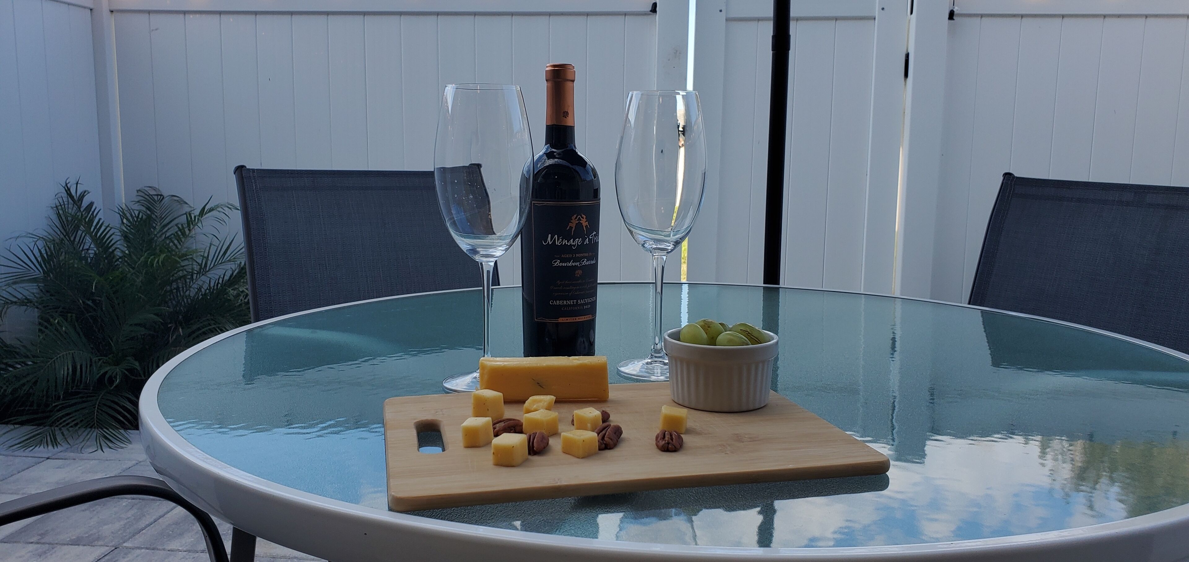 A glass table with a bottle of wine, two glasses, a cutting board with cheese and nuts, and a bowl of olives.