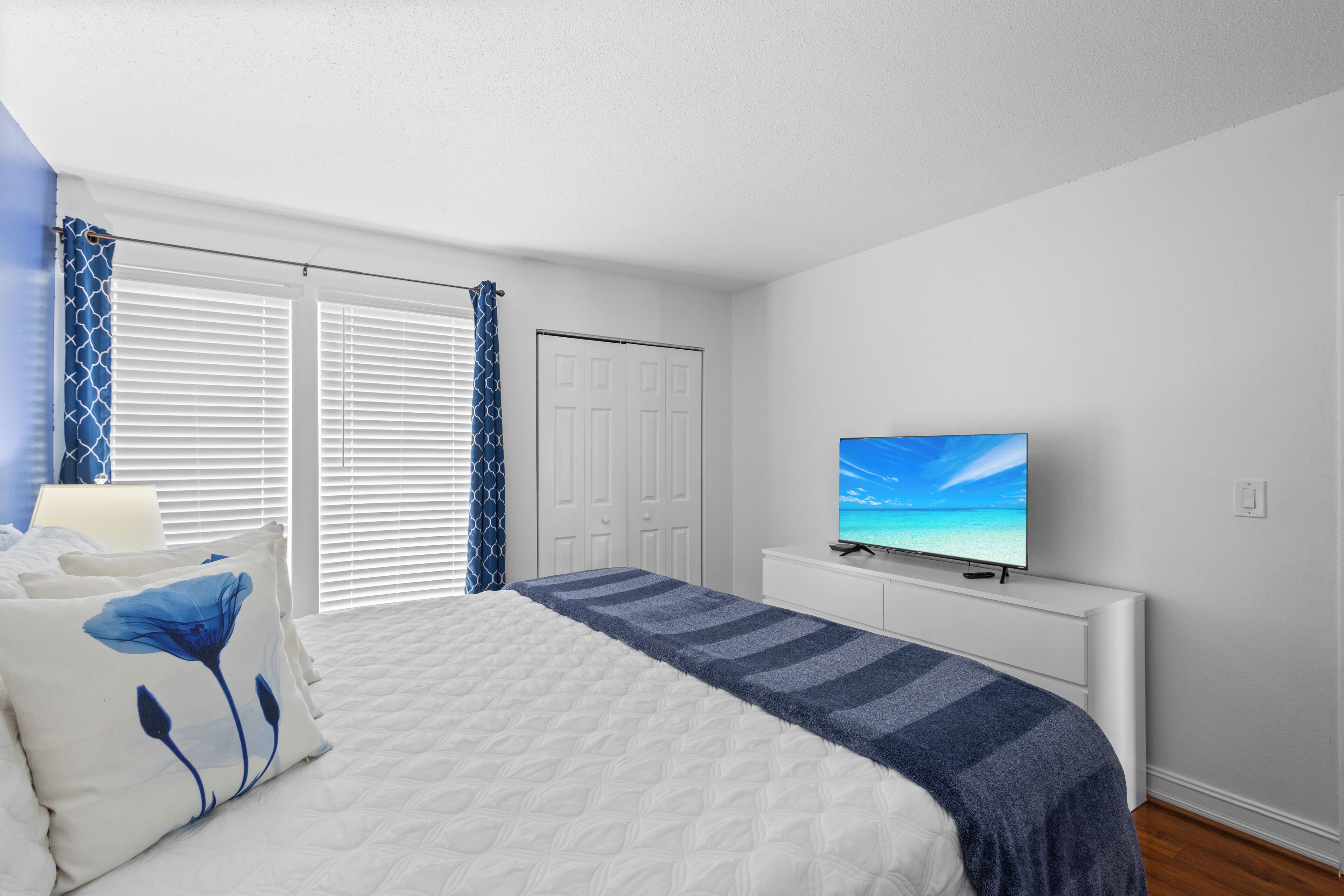 A bedroom with a bed, a television on a stand, a window with blinds, and a door.