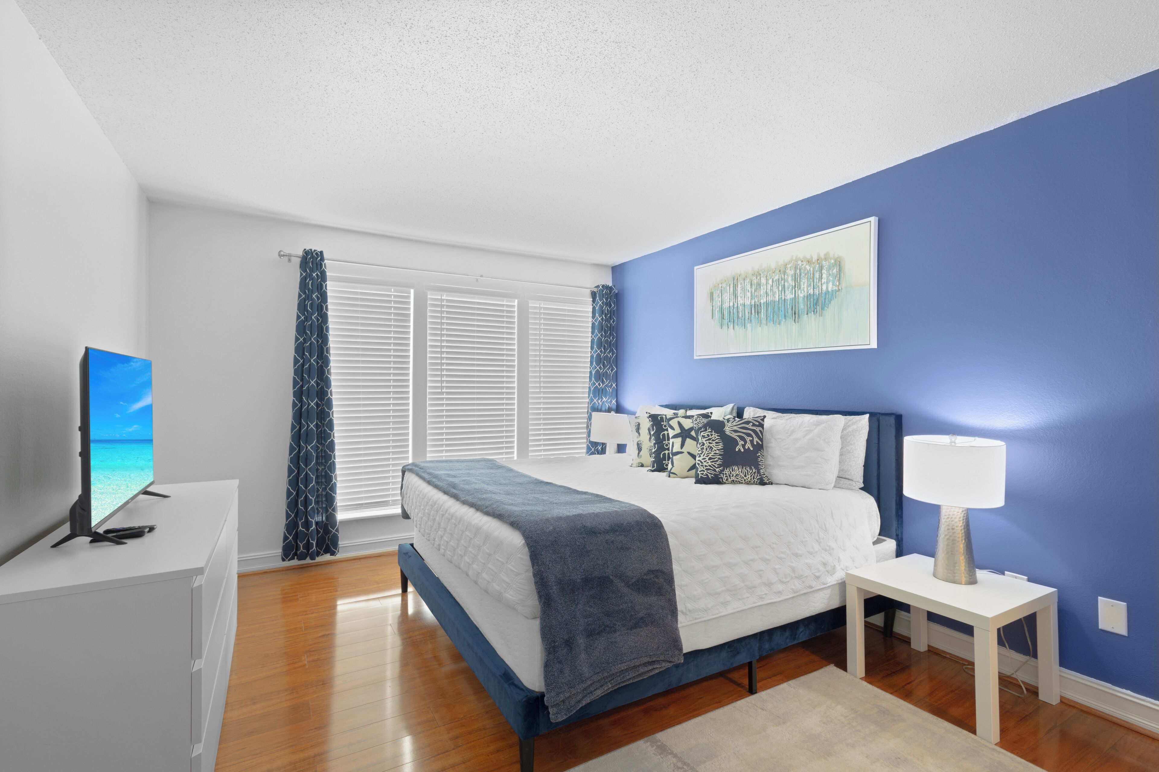 A bedroom with a bed, a bedside table with a lamp, a TV, and a window with blinds.