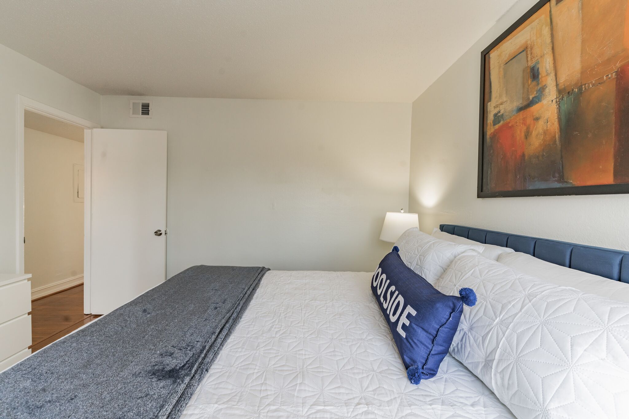 Un dormitorio con cama, almohada azul, colcha blanca acolchada, manta gris y una pintura en la pared, además de una puerta blanca.
