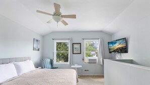 Et soveværelse med en seng, ventilator, fjernsyn og to vinduer med gardiner.