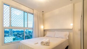 A hotel room with a large window, a bed with white linens, and a remote control on the bed. - Apart em Ponta Negra por Carpediem (Natal)