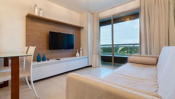 A modern hotel room with a flat-screen TV mounted on a wooden panel above a white console table, a beige sofa, and a glass door leading to a balcony with a view. - Apart em Ponta Negra por Carpediem (Natal)